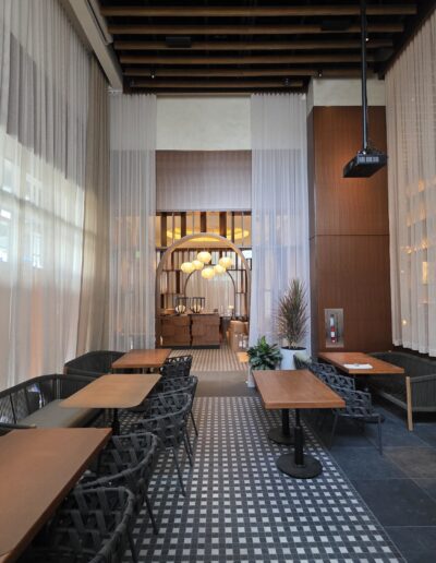 Modern restaurant interior with wooden tables, woven chairs, checkered floor tiles, sheer curtains, and warm lighting; a large archway leads to a seating area in the background.