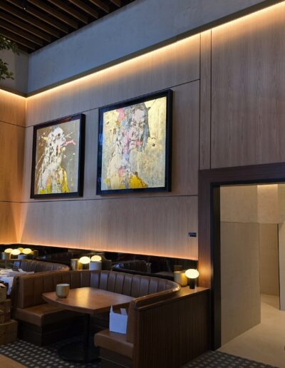 A modern restaurant interior with wooden booths, round tables, wall-mounted lamps, abstract art pieces, and an open doorway leading to another room.