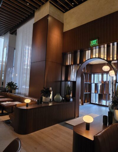 Modern restaurant interior with wooden accents, round tables, booth seating, decorative plants, globe lamps, and large windows letting in natural light.