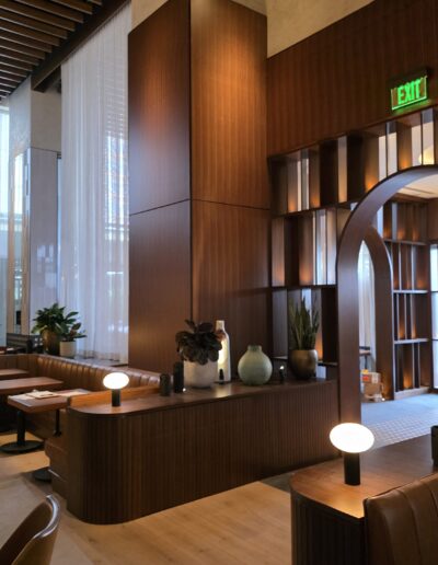 Modern restaurant interior with wooden panels, leather seating, round tables, decorative plants, soft lighting, and an illuminated exit sign.