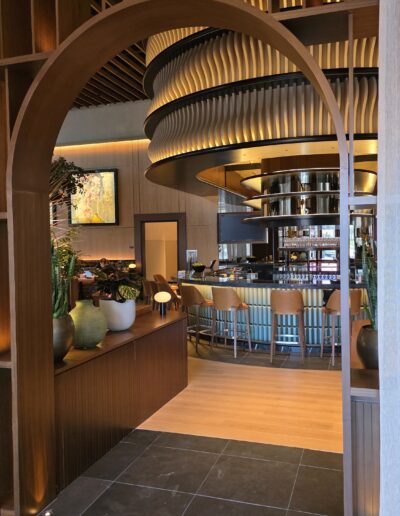 View through a wooden archway into a modern restaurant with curved ceiling details, a central bar area, bar stools, and decorative plants in the foreground.
