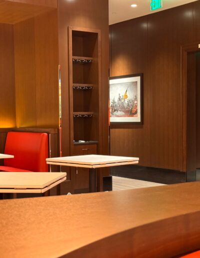 A restaurant interior with red booth seating, wooden tables, and brown walls. A hallway with a restroom sign and a framed picture are visible in the background.