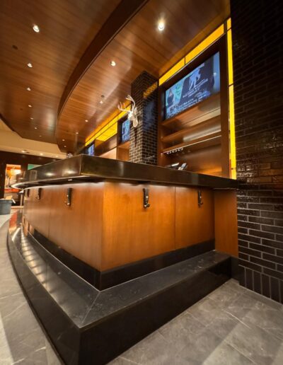 Reception desk with wood paneling, black countertop, and mounted screens above. Overhead lights and a decorative antler mount are visible, with dark brick accents on the right.