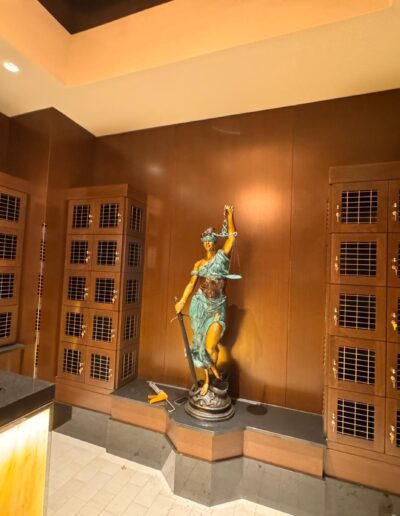 A bronze statue of Lady Justice stands in the corner of a room with dark wood paneled walls and multiple wooden lockers with metal grates.