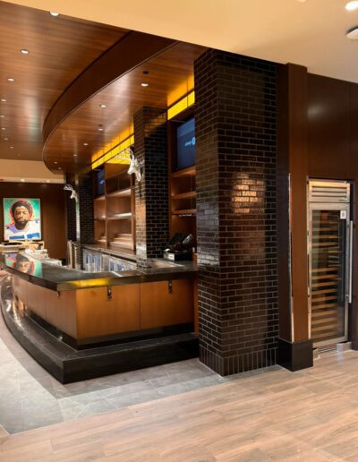 Modern, empty bar area with wooden panels, black brick columns, metal bar counter, and open shelving; a portrait hangs on the back wall.