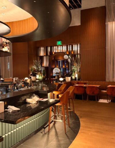 A modern restaurant interior features a marble bar with leather stools, wooden paneling, soft lighting, and decorative plants.