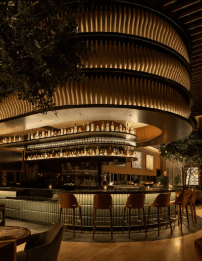 A modern, dimly lit bar with curved shelving stocked with bottles, surrounded by bar stools, plants, and warm ambient lighting.